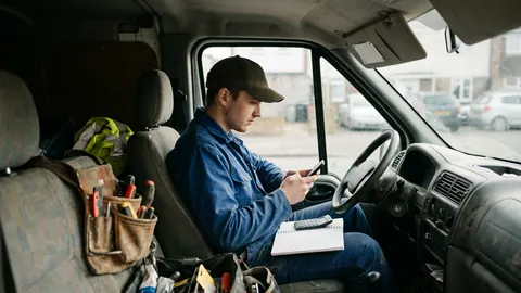 Young tradesman in a work cap sitting in the driver seat of a work van with a notebook and phone, tool belt on the passenger seat